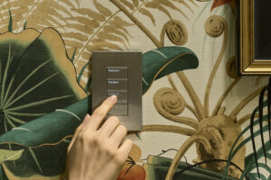 A hand presses a button on a silver Lutron Palladiom keypad on a floral wallpapered wall.
