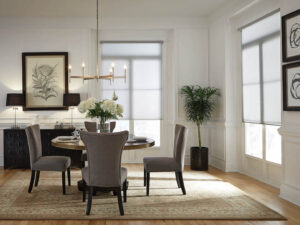 A dining room with a small table, four chairs, and sheer white Lutron roller shades on the windows.